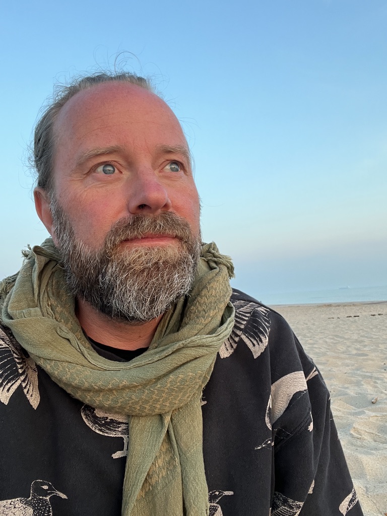 Perry on a beach at dusk, wearing a green scarf and bird-patterned coat